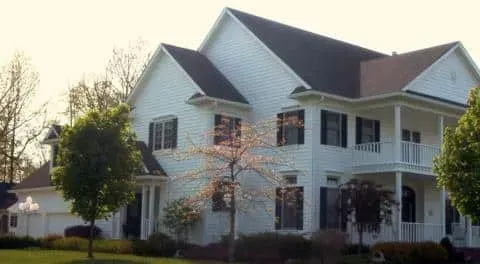 White two-story house with a porch and dark roof, surrounded by trees in daylight.
