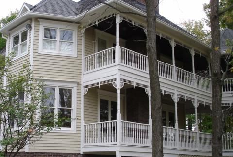 Two-story beige house with white trim and wraparound porches among trees