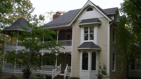 Victorian house with white trim, dark roof, and wraparound porch surrounded by trees