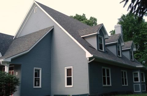 Blue-gray house with steep gabled roof and white trim, shown from the side in a leafy residential setting