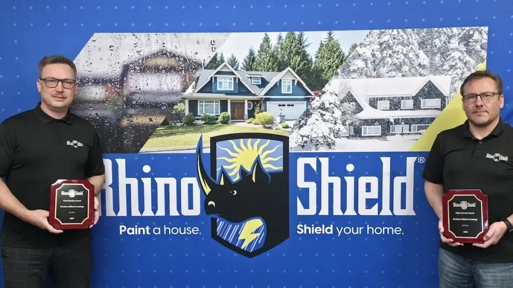 Two men stand beside a Rhino Shield sign and awards in front of a snowy house backdrop.