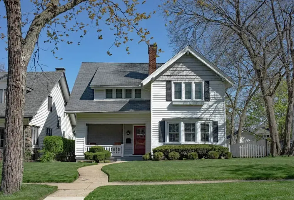 White two-story house with gray roof, front porch, and green lawn on a sunny day