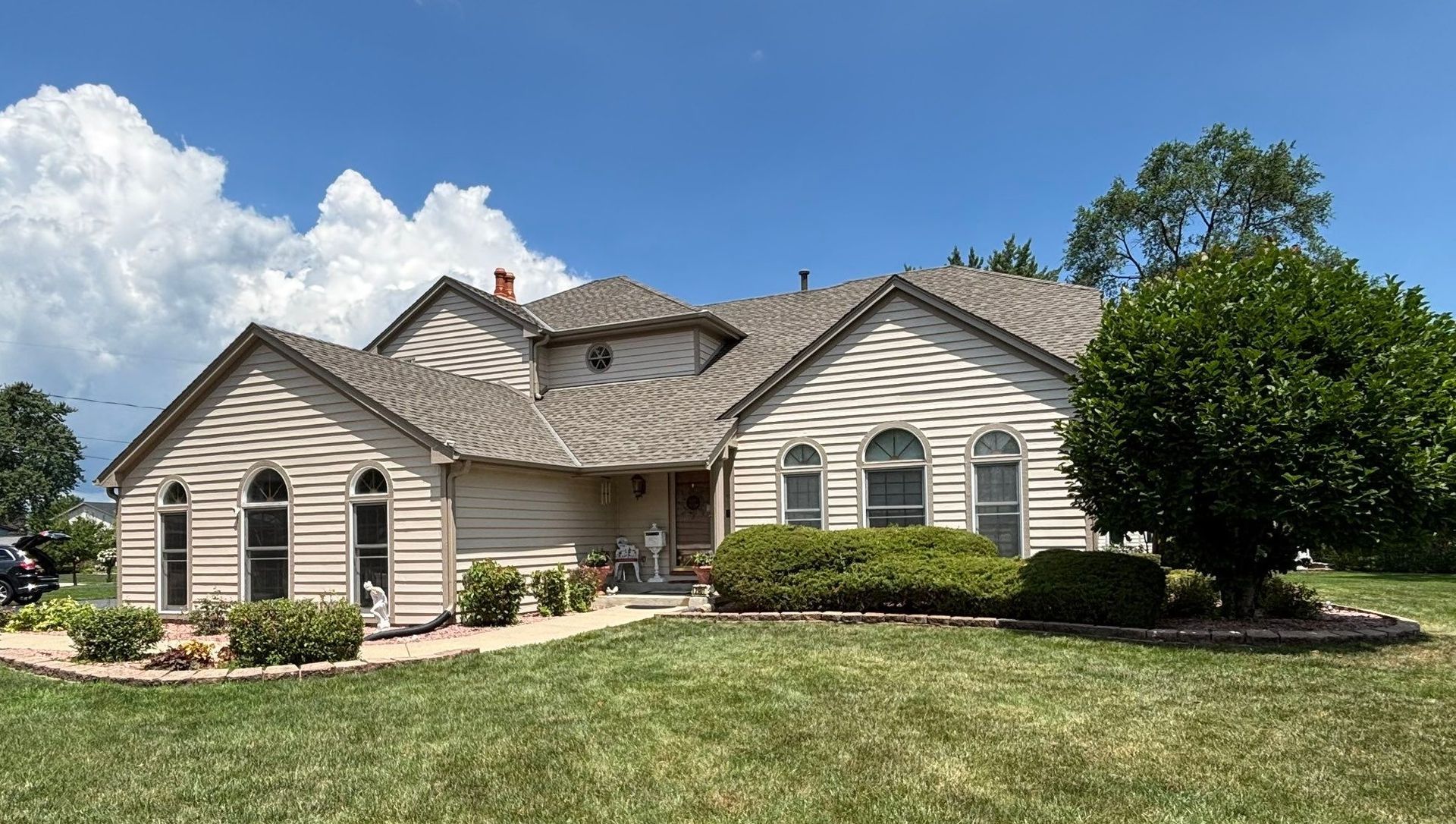Single-story beige suburban house with a manicured lawn, shrubs, and a bright blue sky.