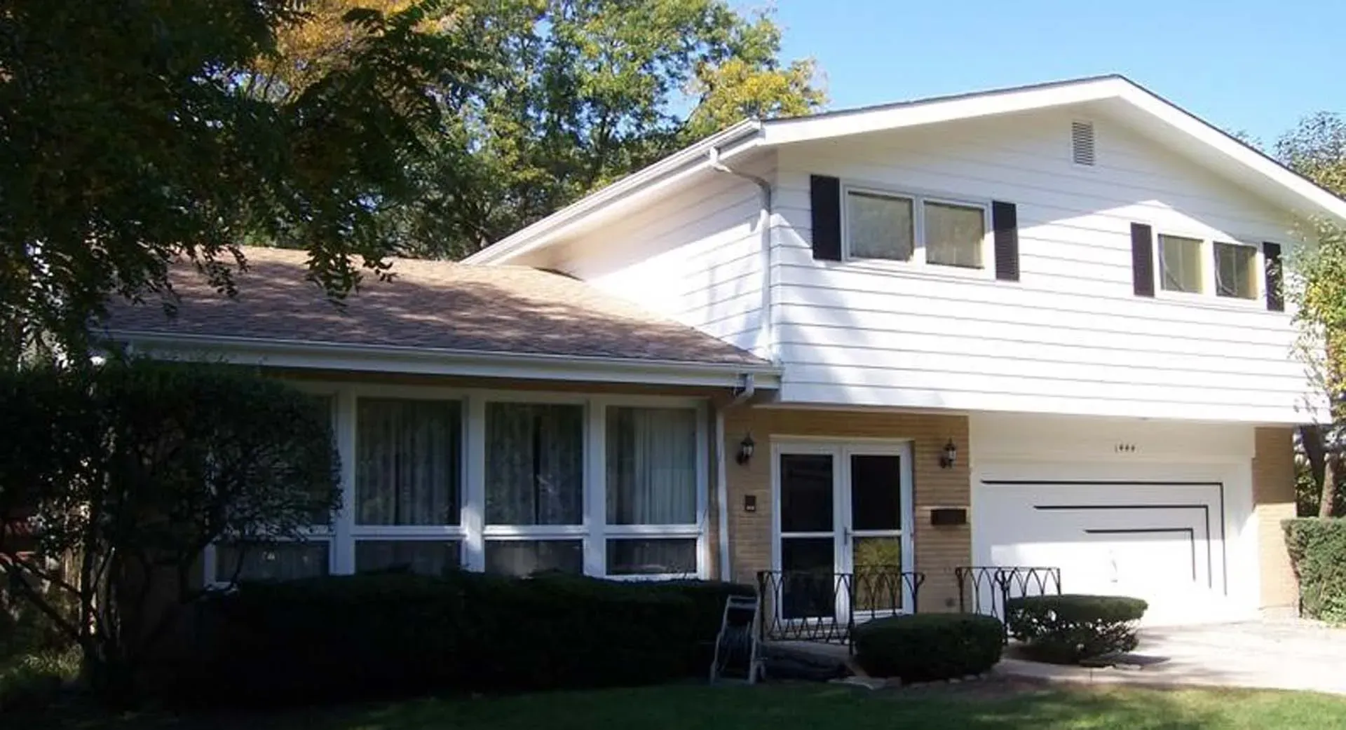 Suburban two-story house with white siding, dark shutters, garage, and front lawn in daylight