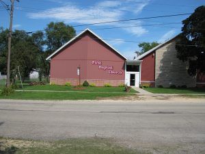 Small pink church building with white trim beside a gray building on a sunny street.