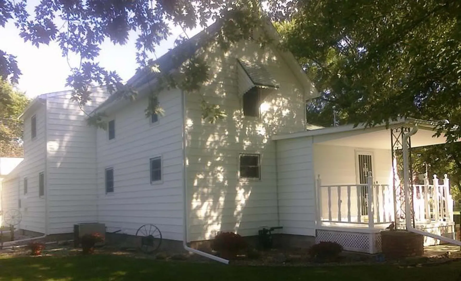 White two-story house with porch, surrounded by trees and sunlight in a leafy yard
