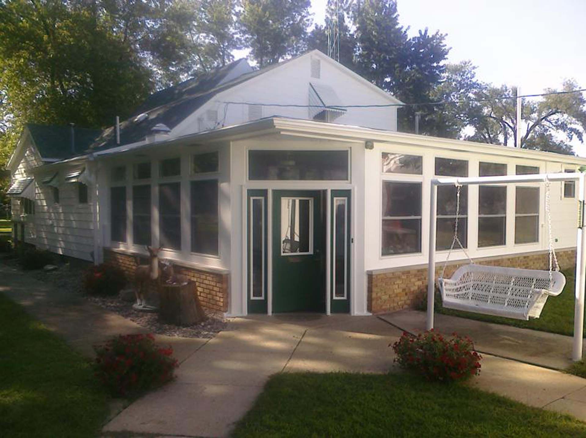 Small white house with a green door, screened porch, and a sign by the walkway under trees.