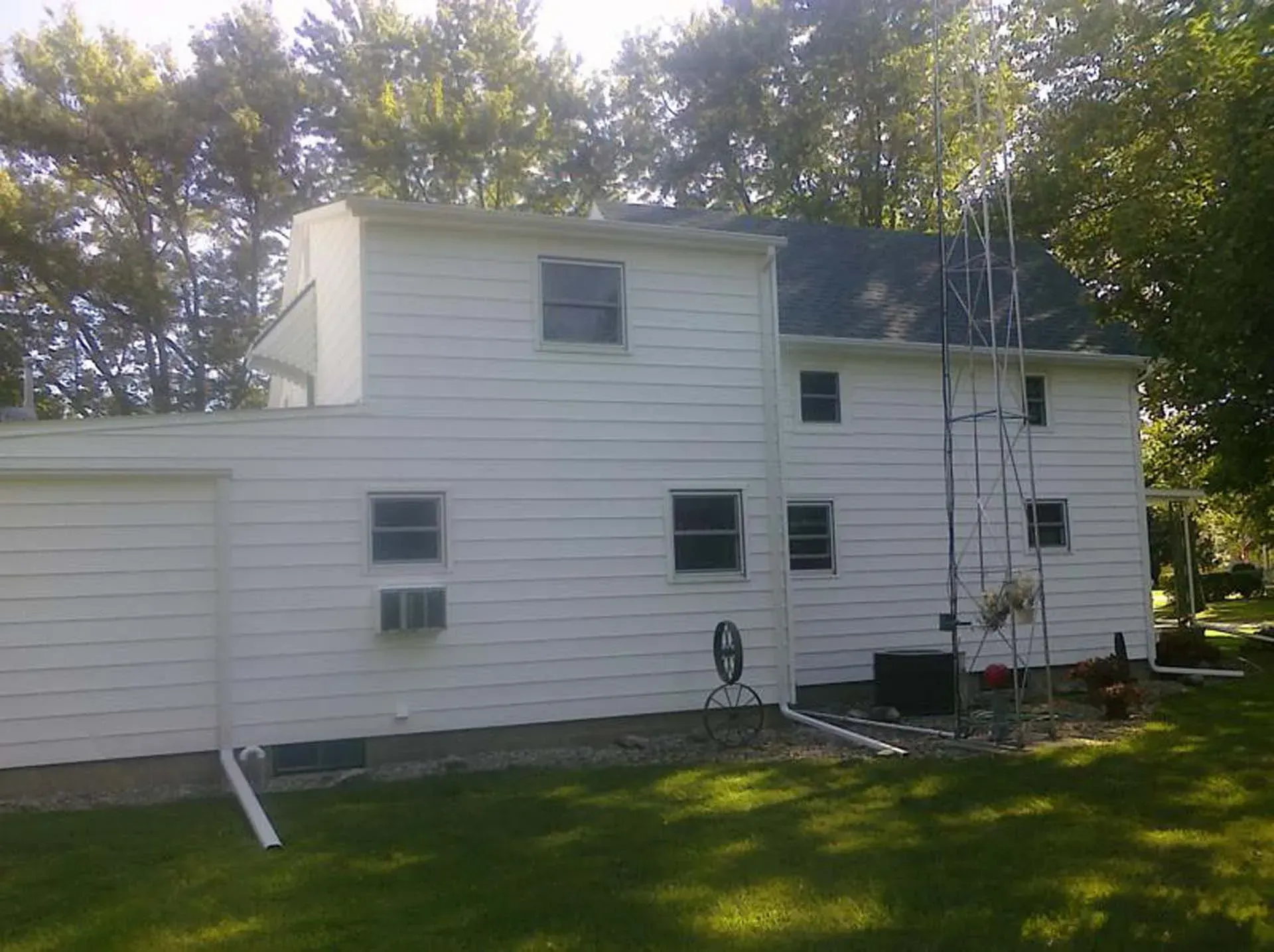 White two-story house with dark roof, small windows, and trees in the background on a sunny day