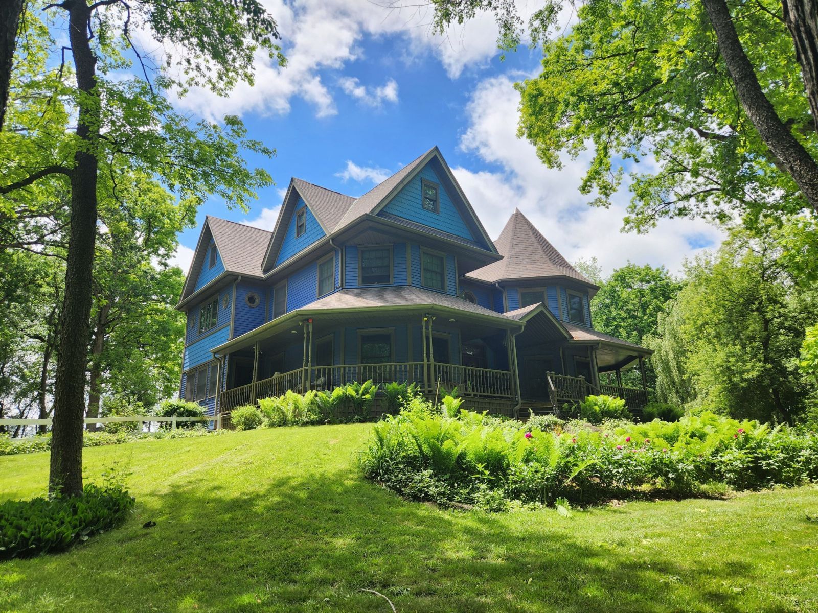 Blue Victorian house with wraparound porch amid green trees and lawn under a partly cloudy sky