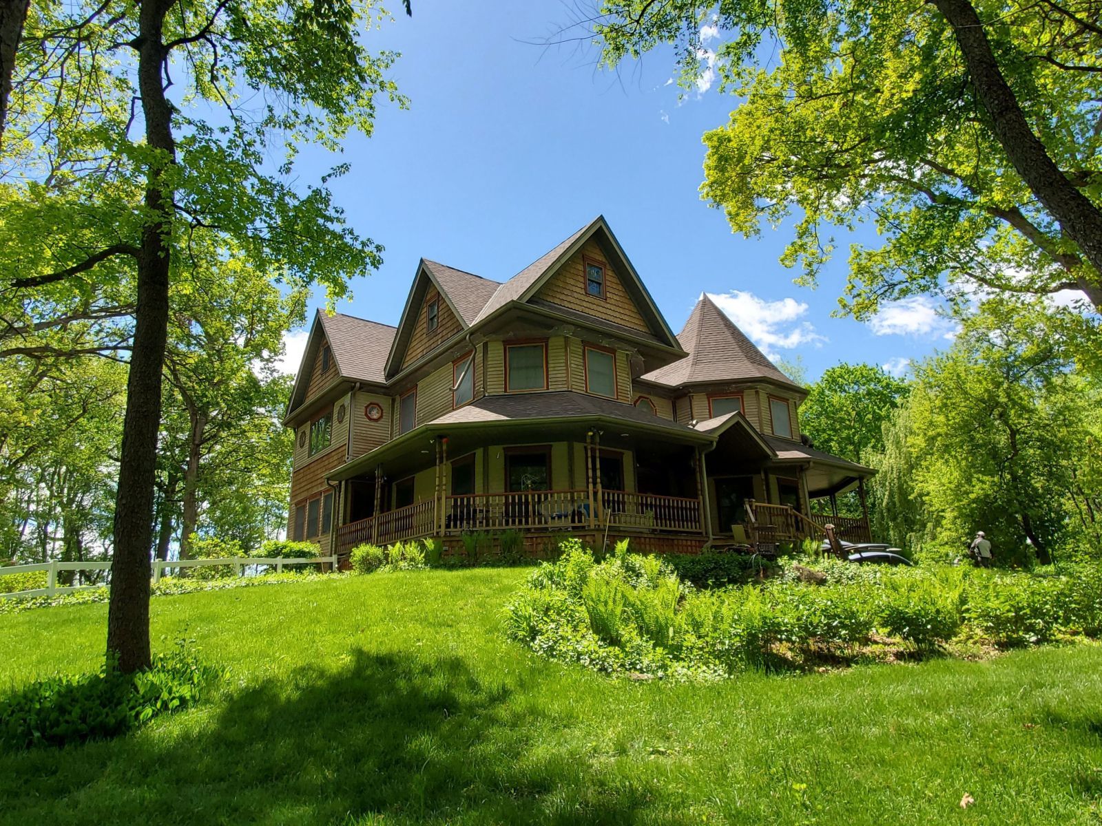 Victorian-style house with a wraparound porch, surrounded by lush green trees and grass on a sunny day