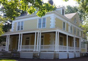 Yellow two-story house with white trim and wraparound porch under trees