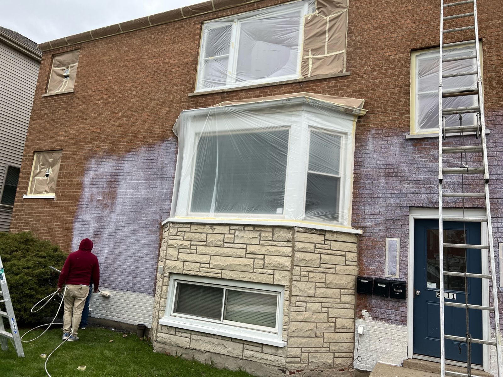 Person painting a brick house exterior beside a ladder and bay window
