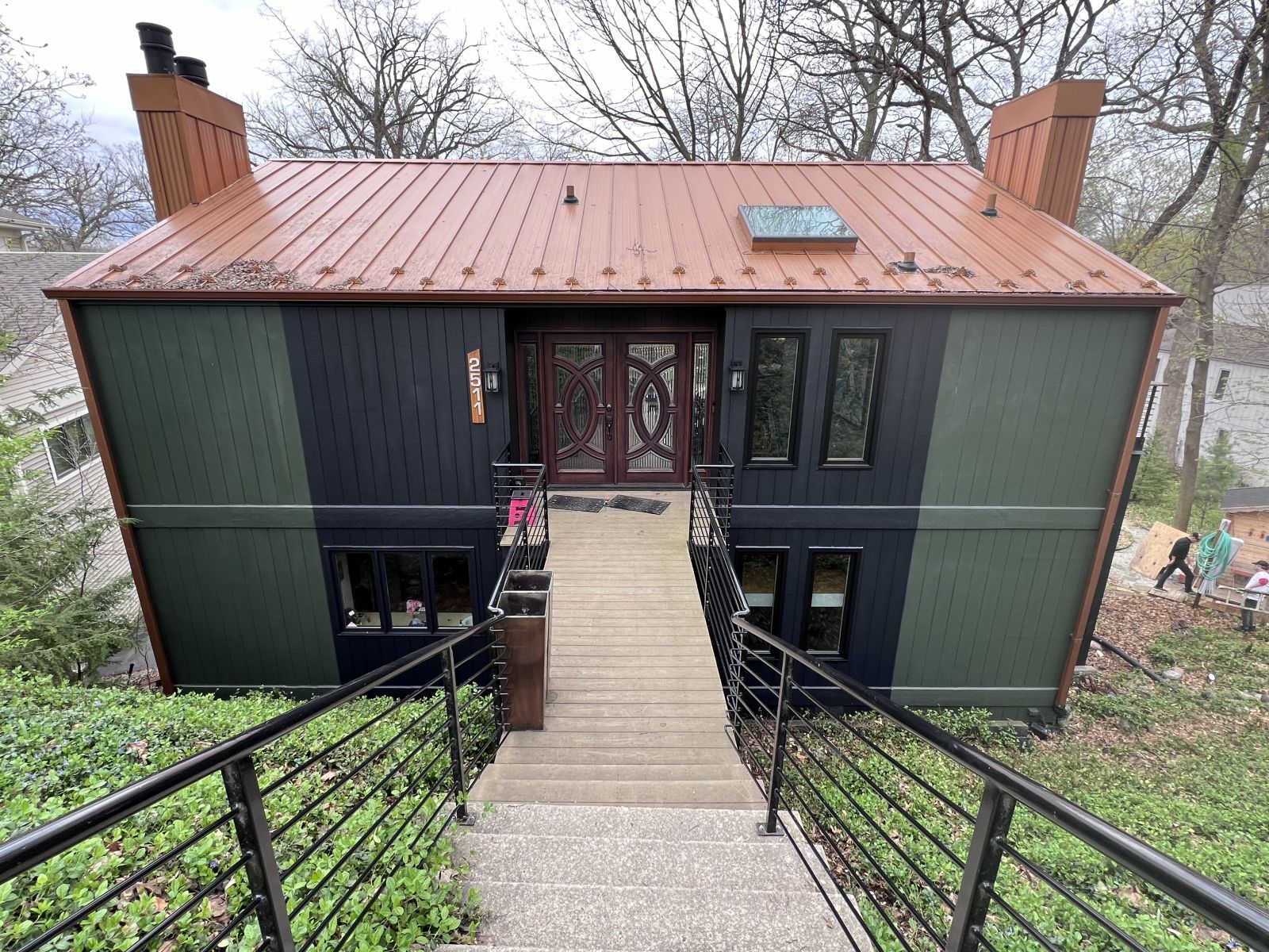 Modern two-story house with copper roof and central ramp leading to the front door, surrounded by trees