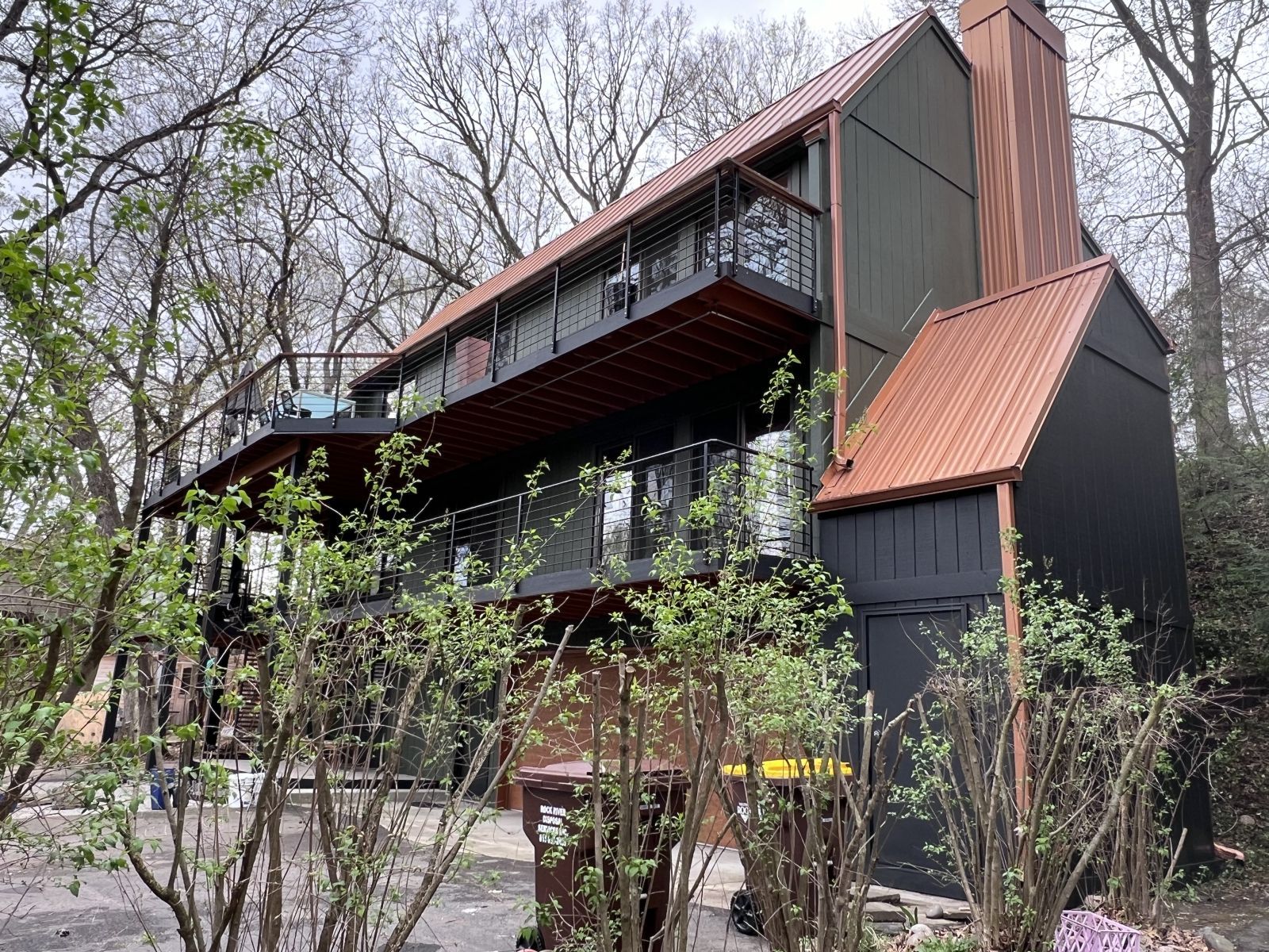 Modern dark house with sloped metal roof on a wooded hillside, surrounded by trees and shrubs.