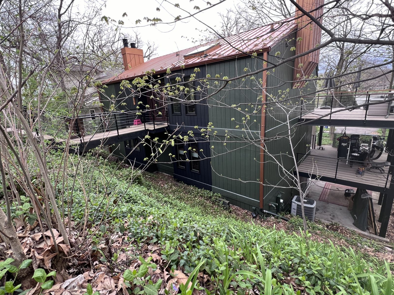 Green house with red roof on a wooded hillside, viewed through bare branches and shrubs