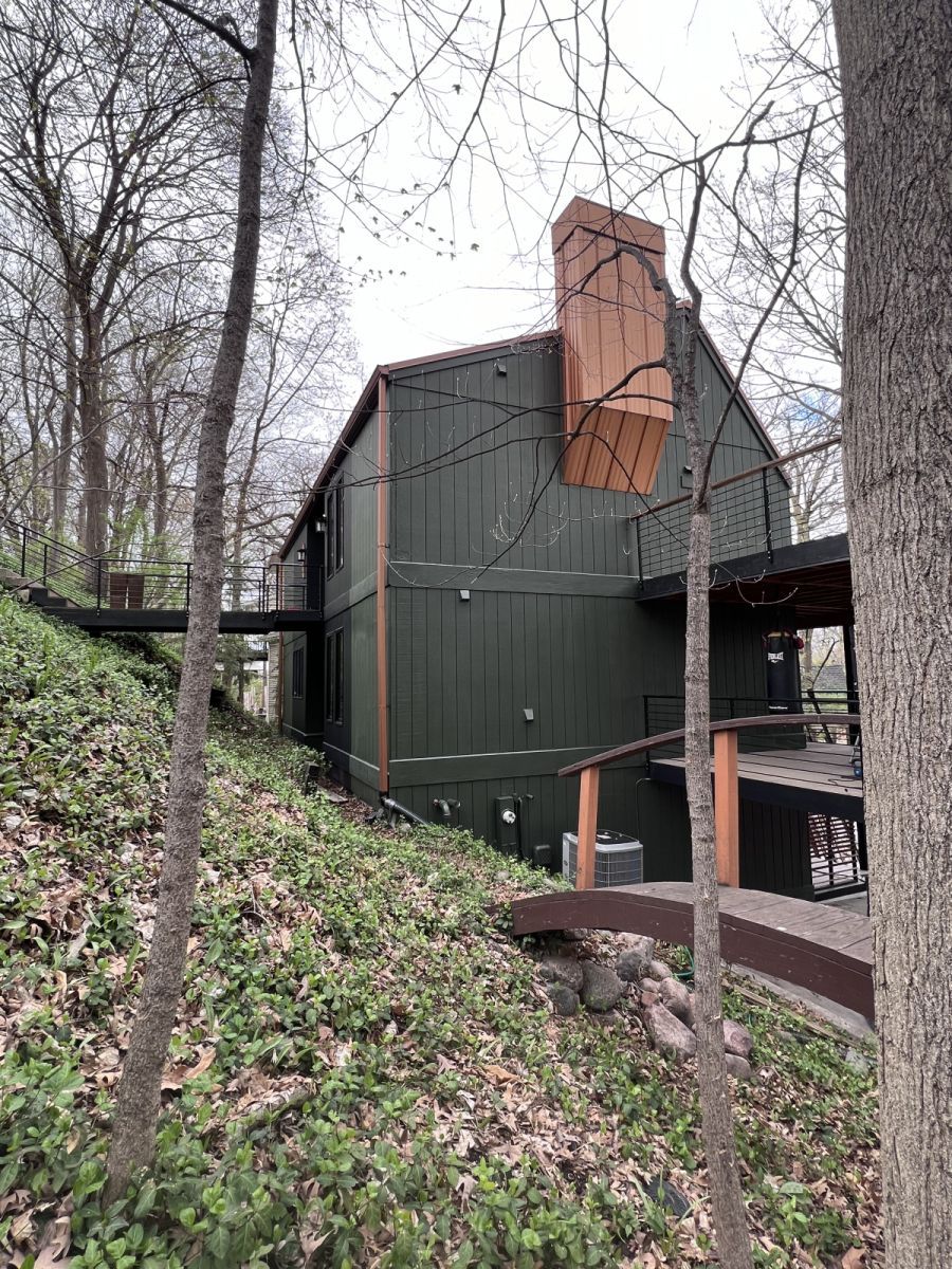 Dark green cabin with red brick chimney on a wooded hillside, viewed from a path.