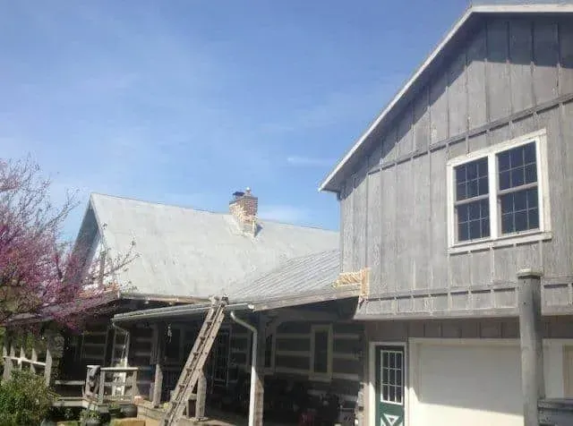 Grey barn-style house with a sloped metal roof, white-trimmed windows, and blooming trees under a blue sky