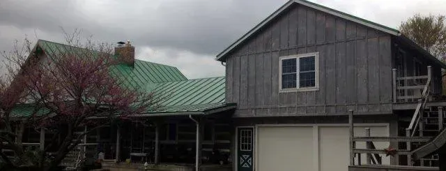 Gray barn with green roof beside a tree and garage under an overcast sky