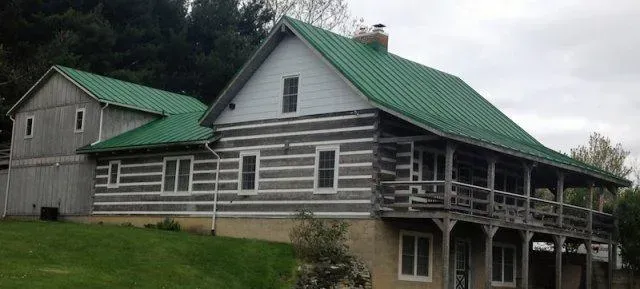 Long rustic log cabin with green metal roofs and wraparound porch on a grassy hillside