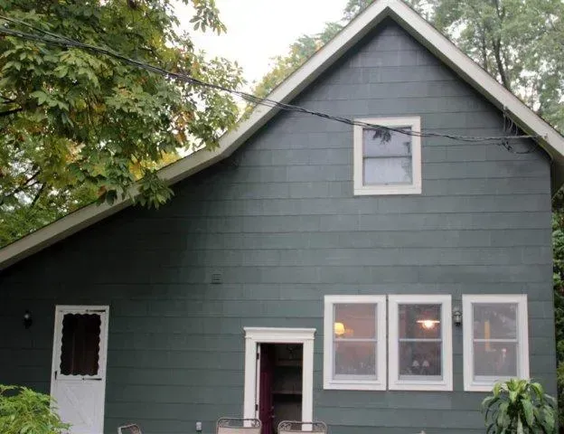 Gray house exterior with white-trimmed windows and a steep roof, partly framed by trees.