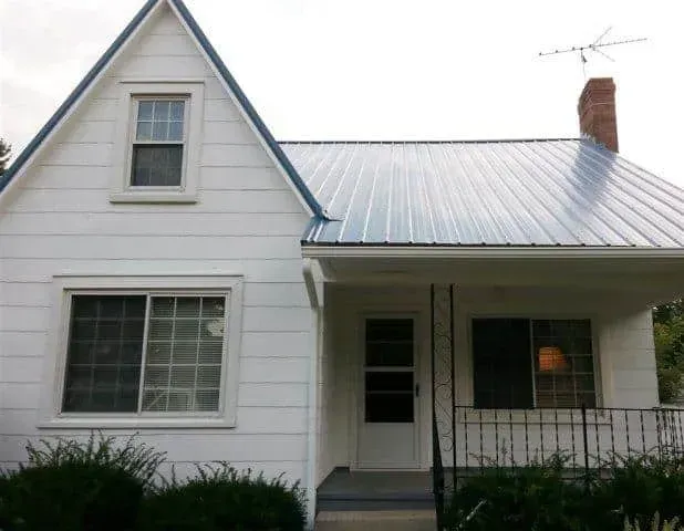 White house with metal roof, front porch, brick chimney, and large windows viewed from the front