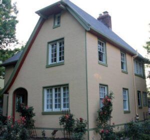 Beige two-story house with green roof and white-trimmed windows, surrounded by shrubs