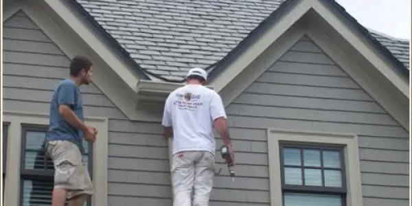 Two men on a roof inspecting gray siding and shingles near a window