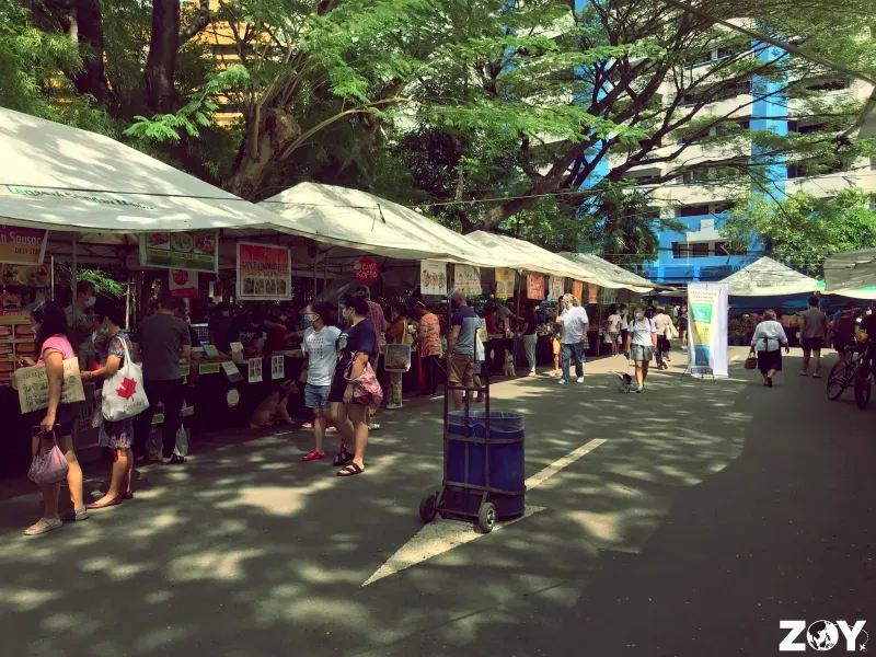 Outdoor market stalls under trees with shoppers walking along a shaded street