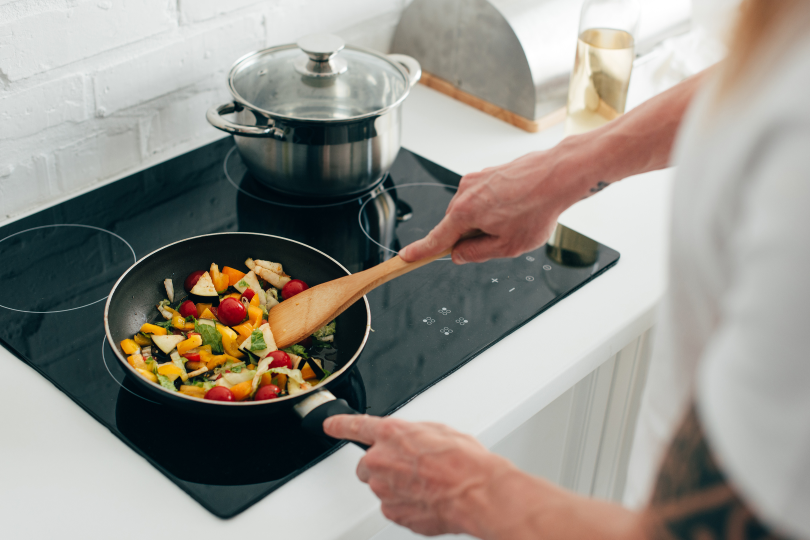 Person cooking vegetables in a pan on a stovetop with a wooden spatula. A pot sits nearby.