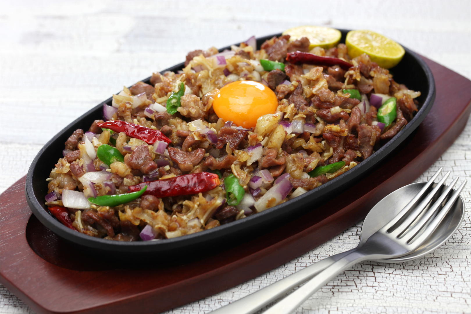 Sizzling Filipino sisig on a dark platter with egg yolk, onions, chilies, and lemon, served on a wooden board.