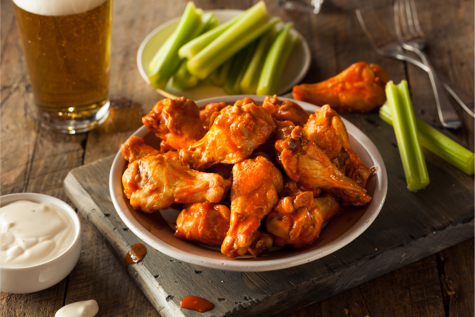 Plate of buffalo wings, celery sticks, ranch dip, and beer on a wooden table.