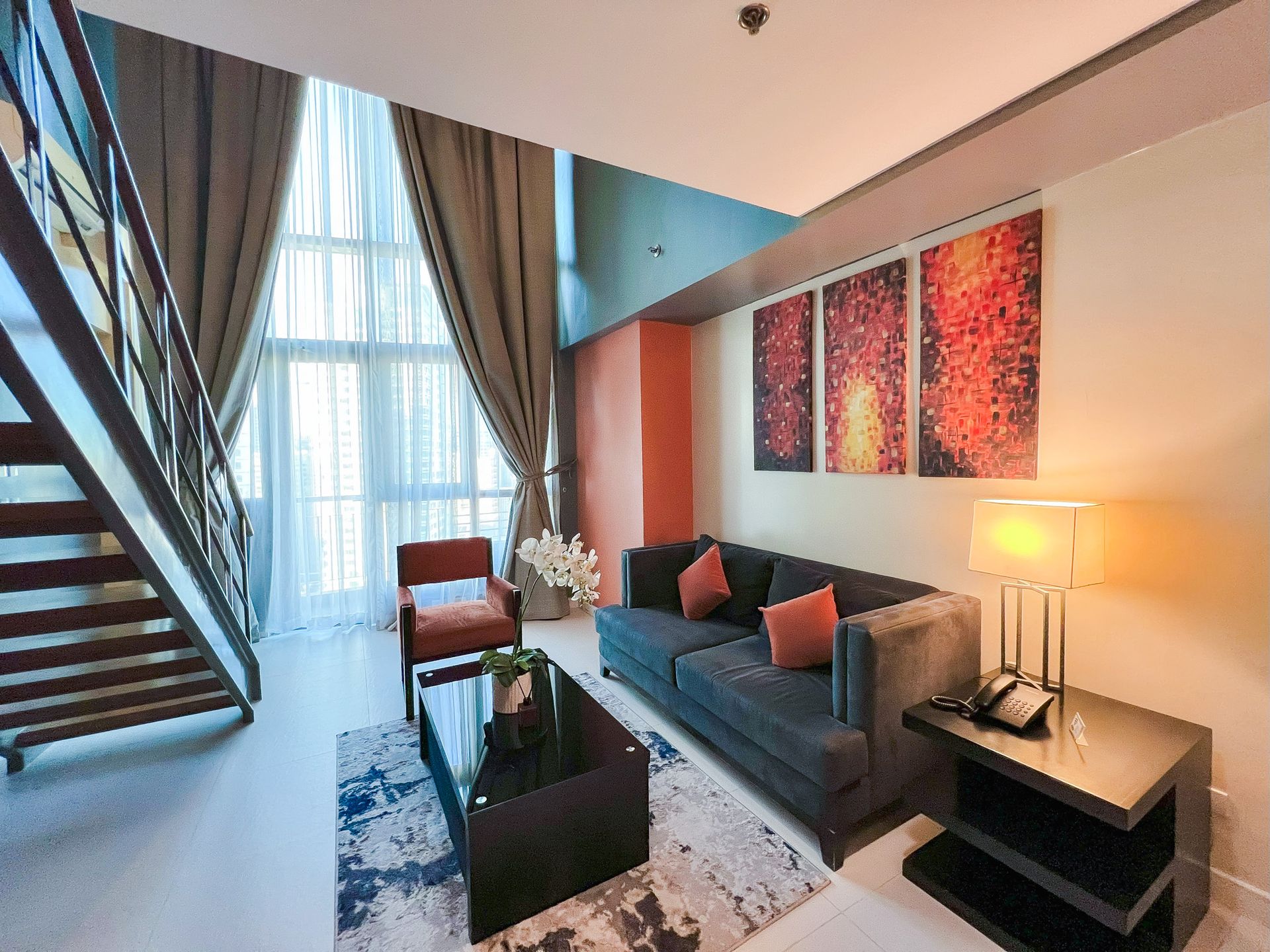 Modern living room with staircase, sofa, coffee table, art, and large windows.