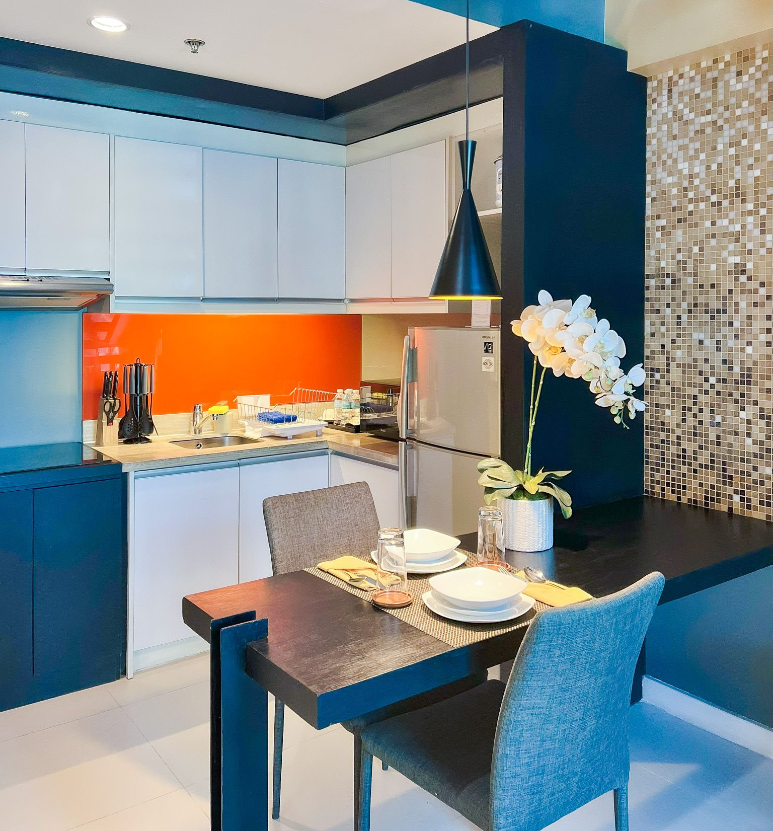 Compact kitchen with dining table: white cabinets, orange backsplash, dark blue accents, and a faux orchid.
