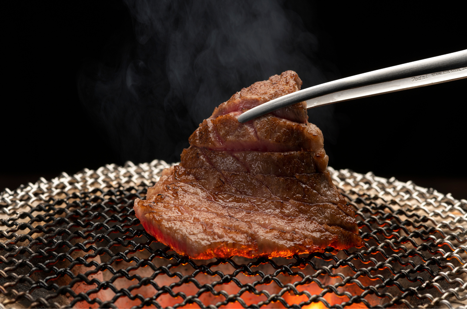 Close-up of a steak being grilled on a metal grate, held by tongs, with smoke rising.