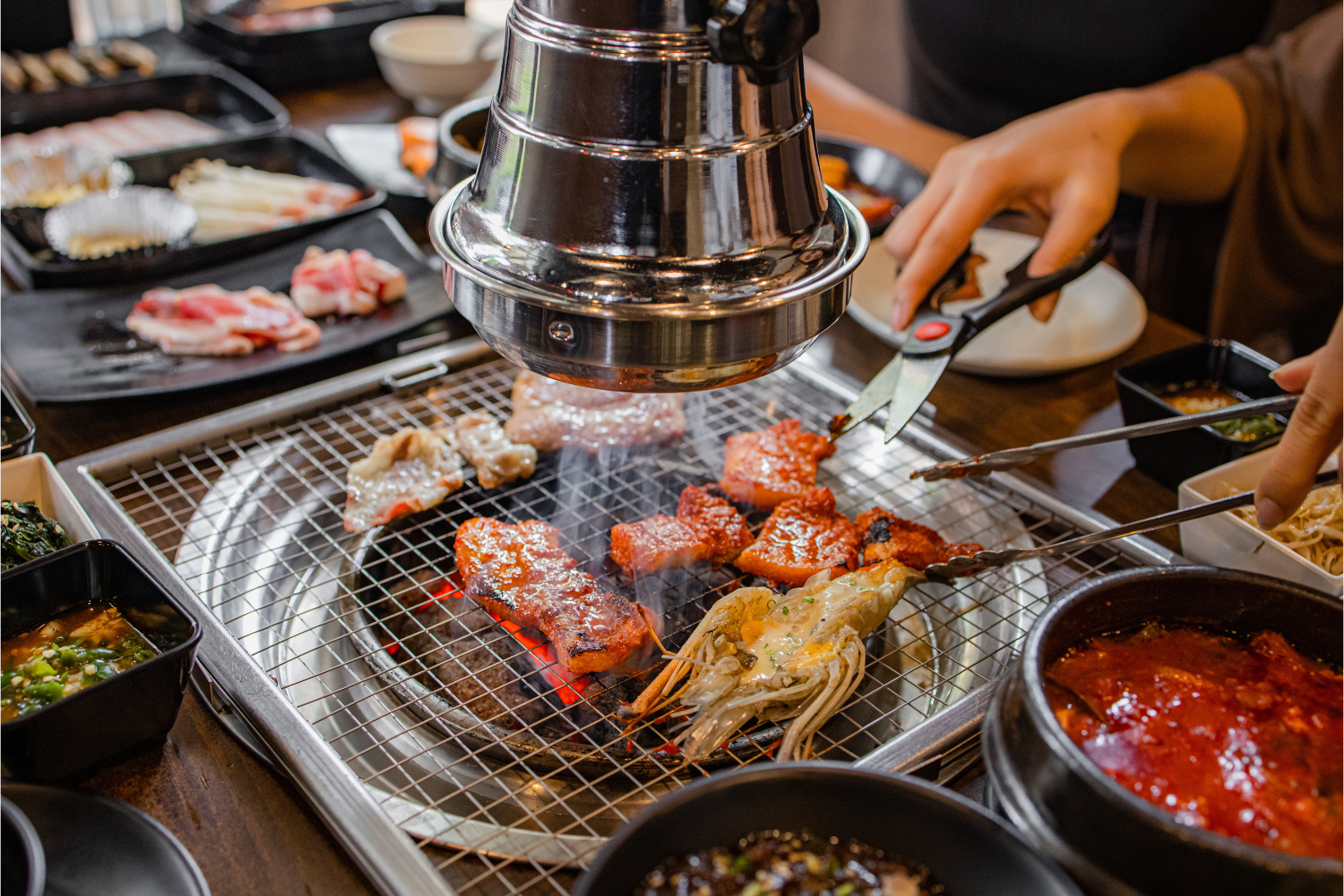 Korean BBQ meal: Meat grilling on a tabletop grill surrounded by side dishes and hands using scissors.