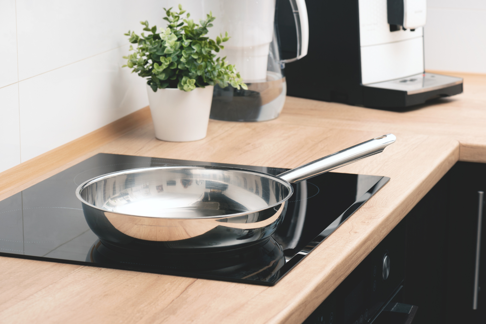 Stainless steel frying pan on a black stovetop in a kitchen setting with a potted plant and coffee machine.