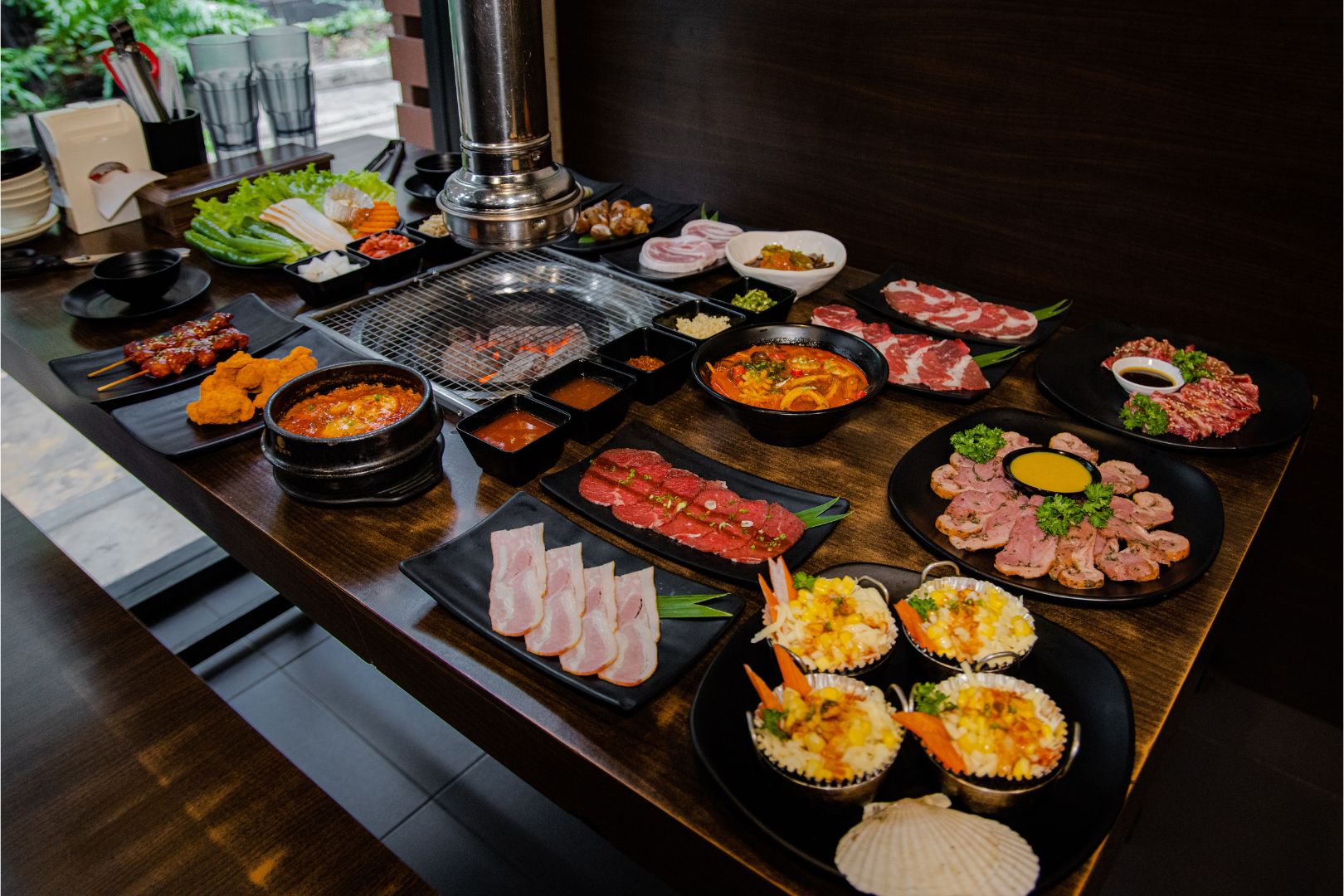 Korean BBQ spread on a dark wooden table with various meats, sides, and a grill.