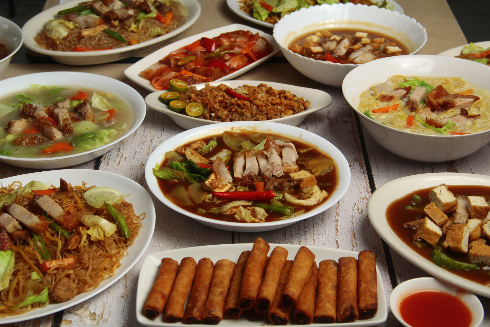 A table spread with various Filipino dishes, including noodles, soup, and spring rolls.