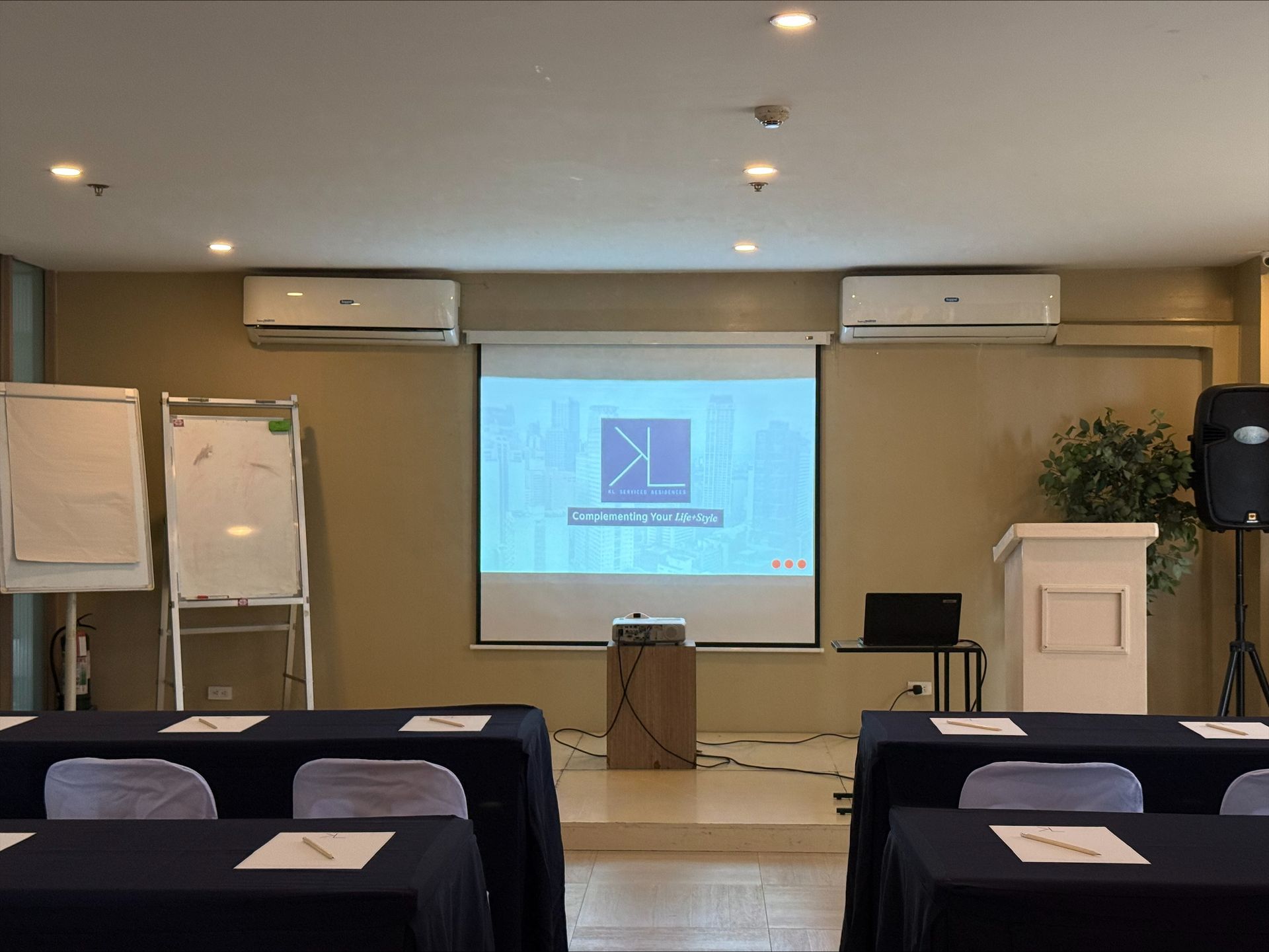 A meeting room with a projector screen, whiteboards, a podium, and rows of tables covered in navy blue cloths.