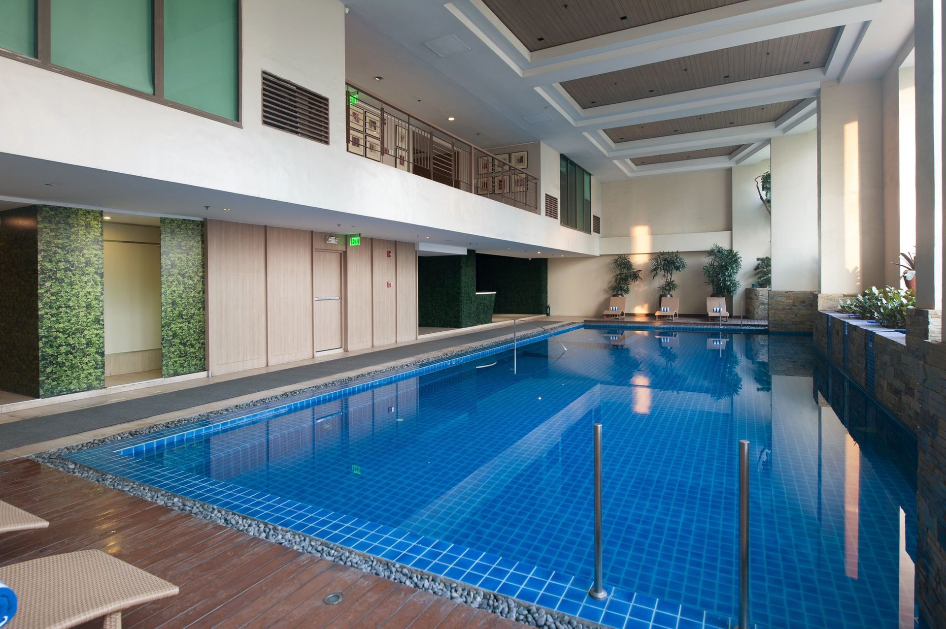 An indoor rectangular swimming pool with blue tiled floor and water, surrounded by wooden decking and a modern building.