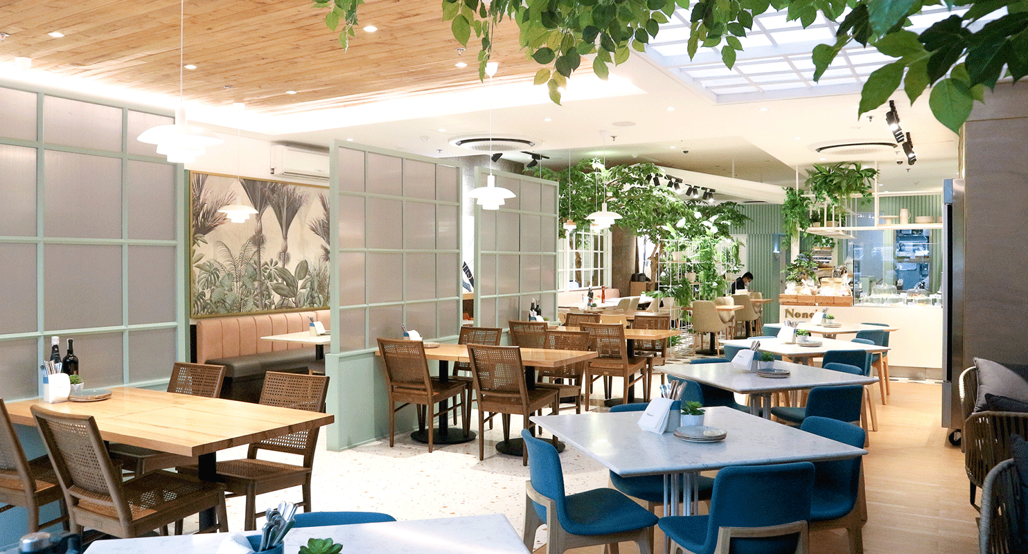 A brightly lit indoor restaurant with wooden tables, rattan chairs, teal accents, and lush hanging greenery.
