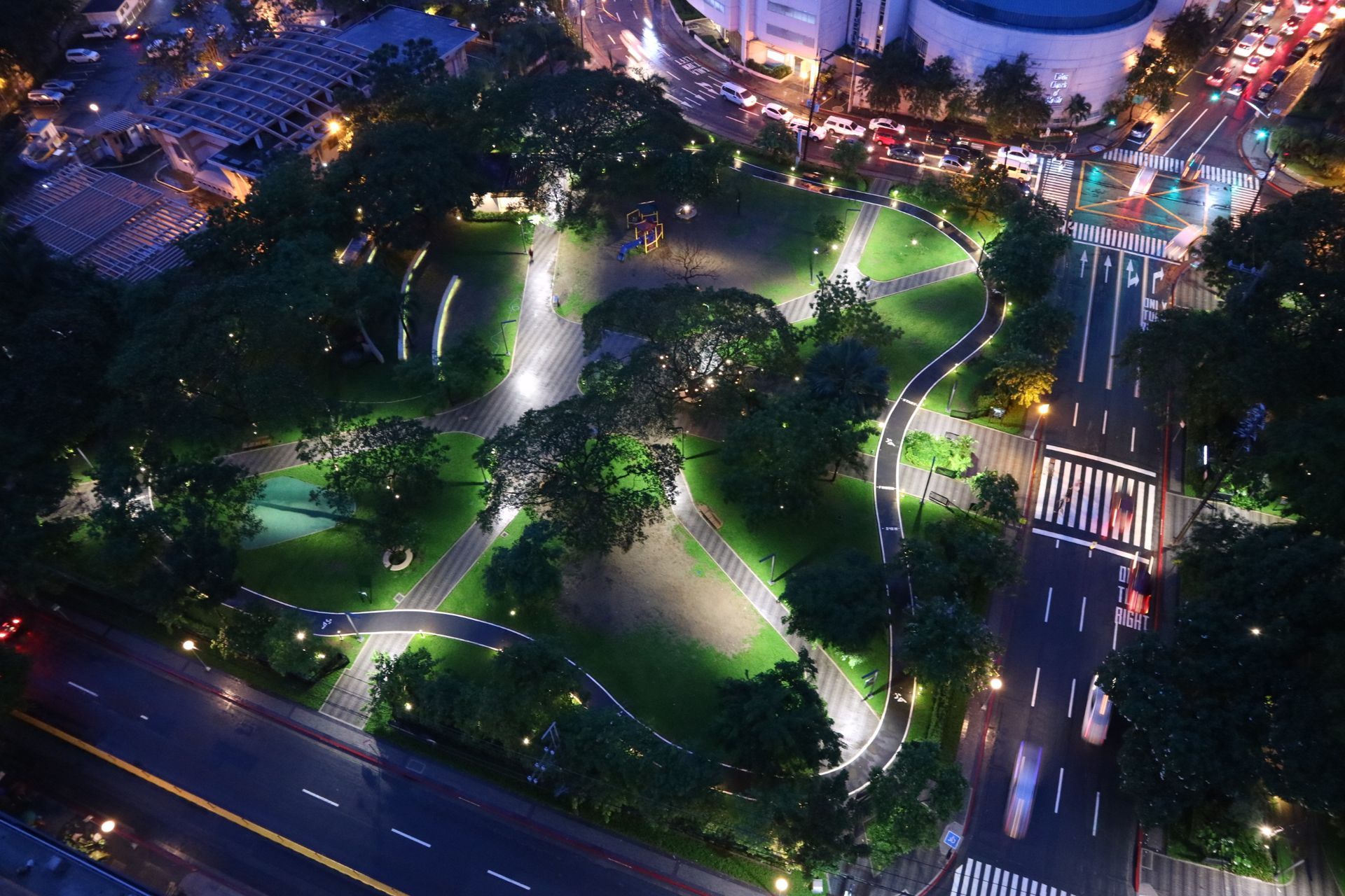 Aerial nighttime view of Legazpi Park in Makati, showing illuminated winding pathways through green lawns and mature trees, a small pond, and a playground, with city streets and traffic surrounding the park