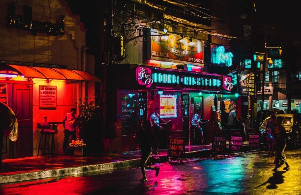 Neon-lit street at night; bar with red and turquoise lights; people walk on a wet, reflective street.