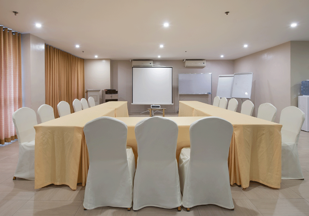 A U-shaped conference table with white-covered chairs arranged in a room with a projector screen and whiteboards.