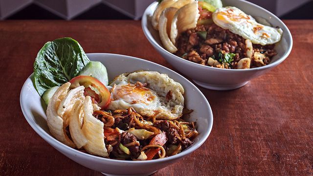 Two bowls of fried rice with fried eggs, crackers, and vegetables on a wood surface.