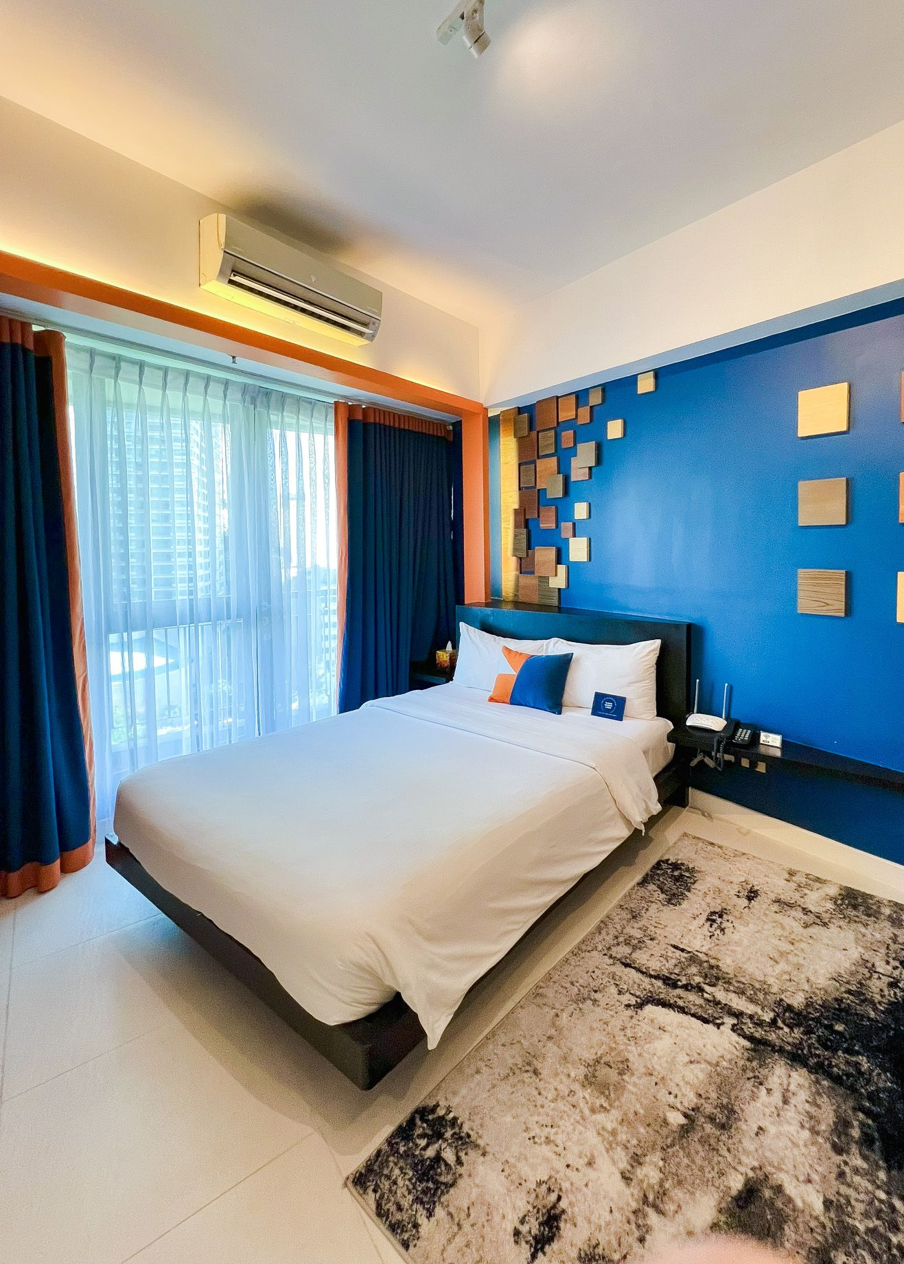 Bedroom with a bed, blue walls, and a patterned rug. Orange and blue curtains frame a balcony.