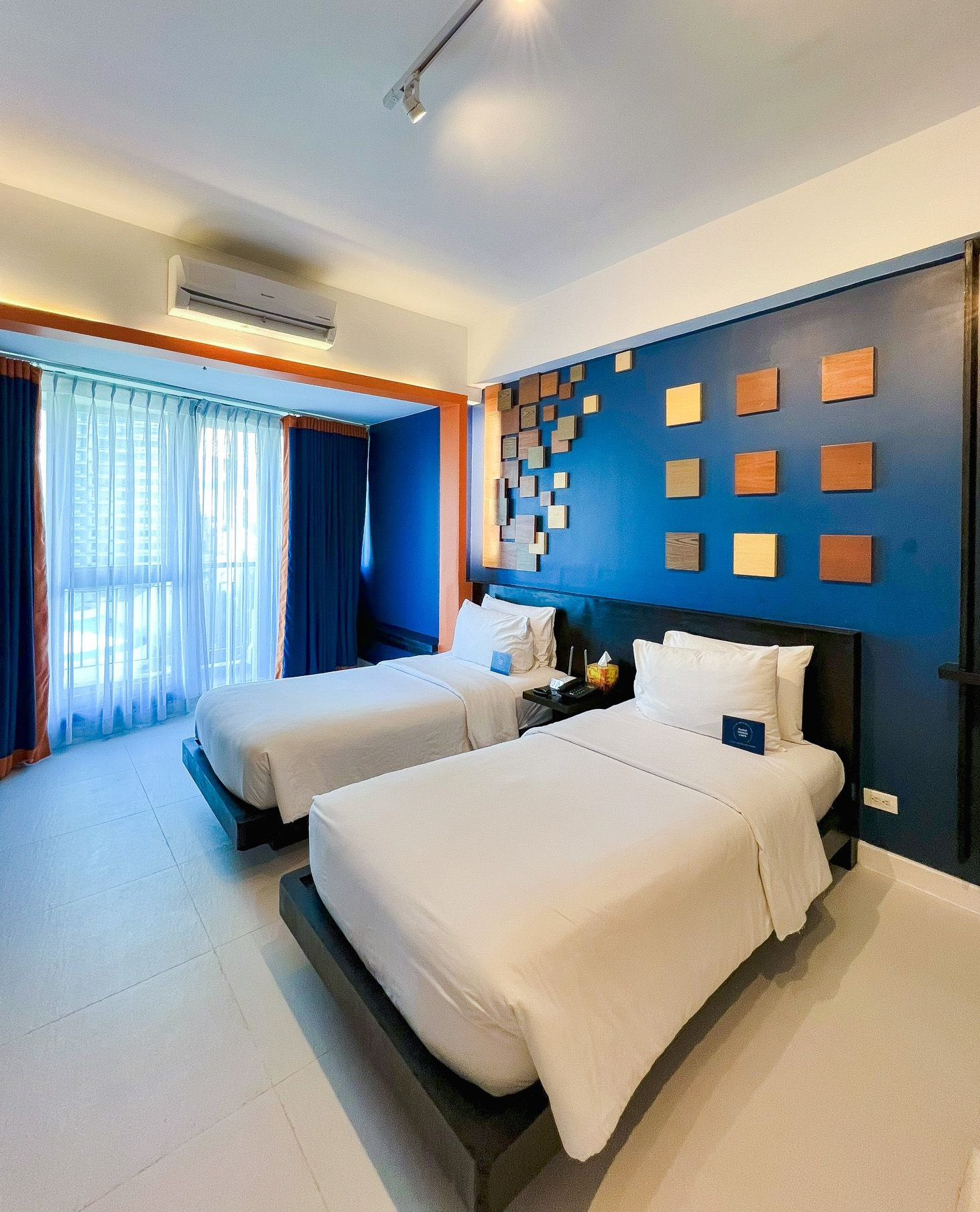 Two twin beds in a hotel room with a blue accent wall and decorative squares.