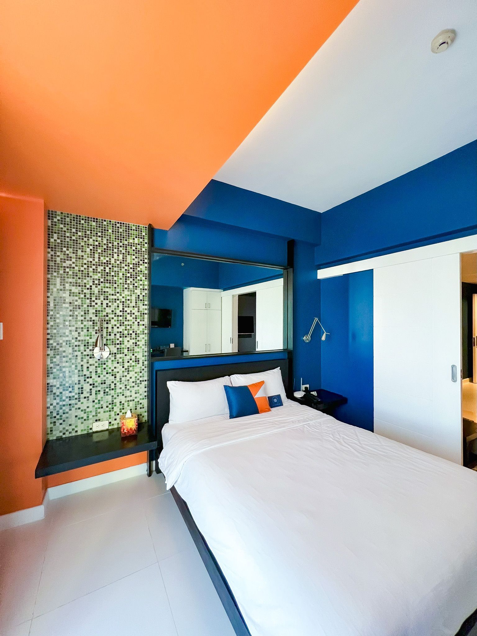 Modern bedroom with orange and blue walls, white bedding, a large mirror, and a sliding door.