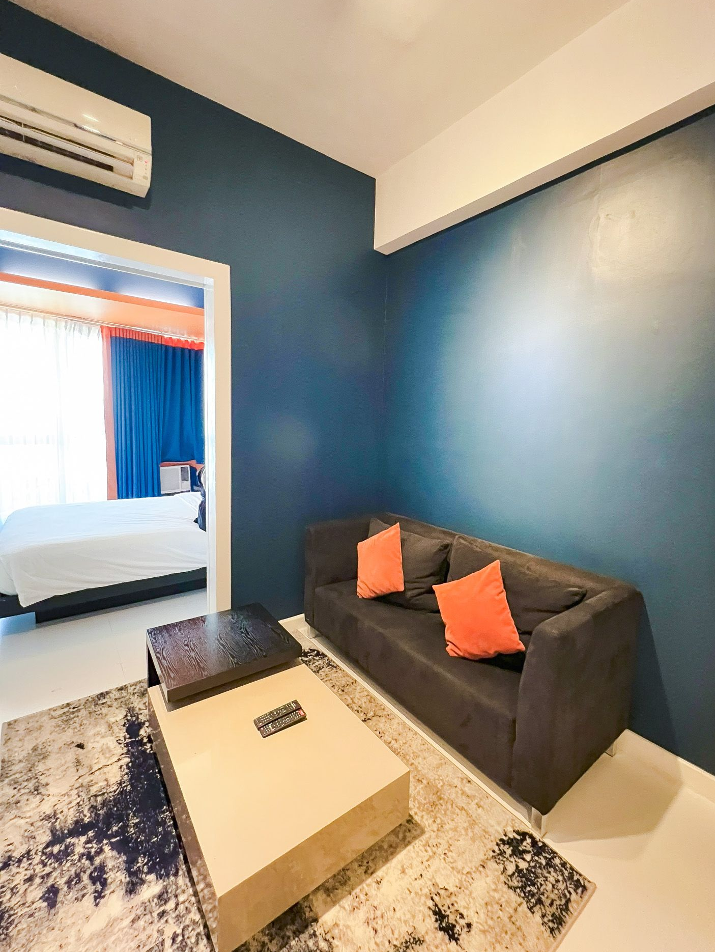 Living room with dark blue wall, gray couch, orange pillows, and a view into a bedroom.