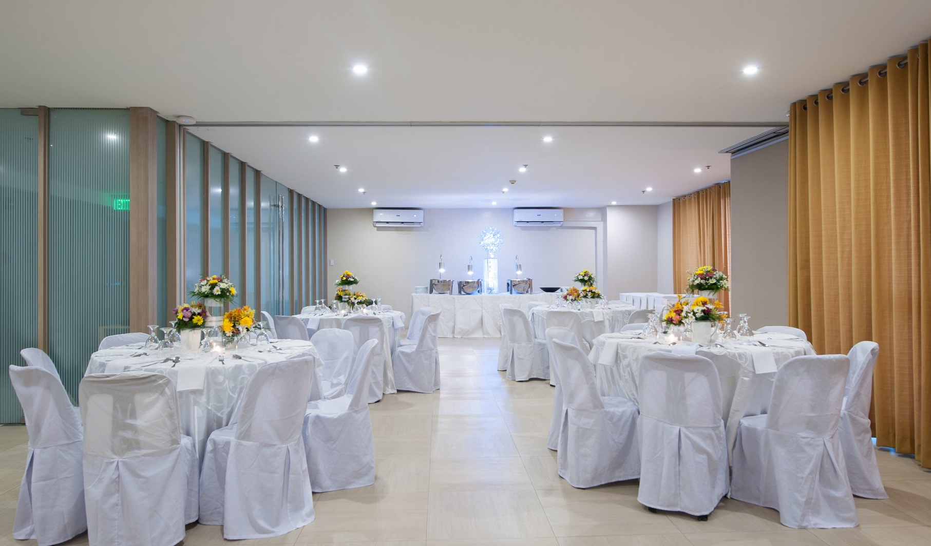 A banquet hall with tables set for a formal event. White linens, flowers, and drapery are featured.